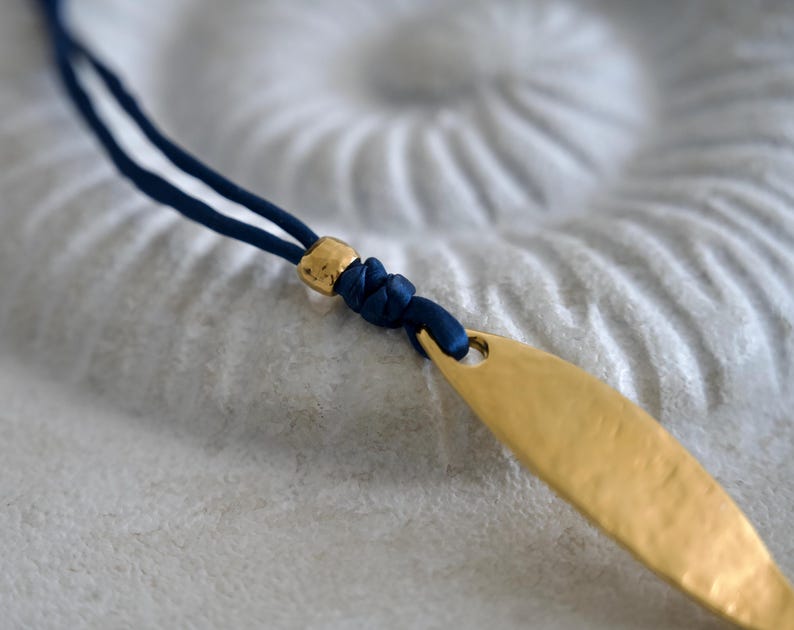 May include: A necklace featuring a dark blue cord, a small metallic gold bead, and a textured, elongated gold-toned pendant. The cord is knotted securely through the top of the pendant, creating a simple design with contrasting blue and gold colors.