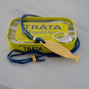 May include: A gold-toned fish-shaped pendant on a blue cord necklace with two small gold-toned square beads featuring a Greek key pattern. The necklace is resting on a yellow rectangular tin of Trata sardines with a silver pull-tab lid.