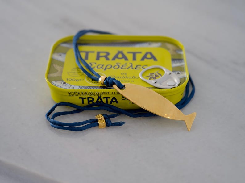 May include: A gold-toned fish-shaped pendant on a blue cord necklace with two small gold-toned square beads featuring a Greek key pattern. The necklace is resting on a yellow rectangular tin of Trata sardines with a silver pull-tab lid.