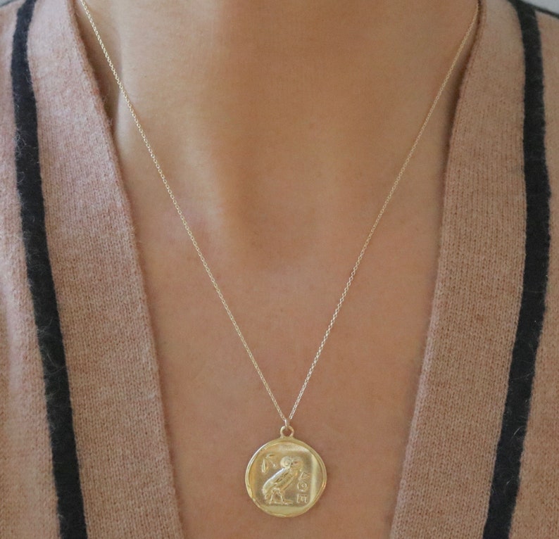 Op de afbeelding: Een goudkleurige ketting met een ronde hanger met een uil en Griekse letters. De hanger hangt aan een fijne ketting. De ketting wordt gedragen tegen een neutraal gekleurde trui.