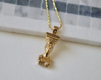 May include: Gold-colored pendant necklace featuring a classical bust design. The pendant is intricately detailed and suspended from a delicate gold chain. The necklace is displayed on a white and gray surface.