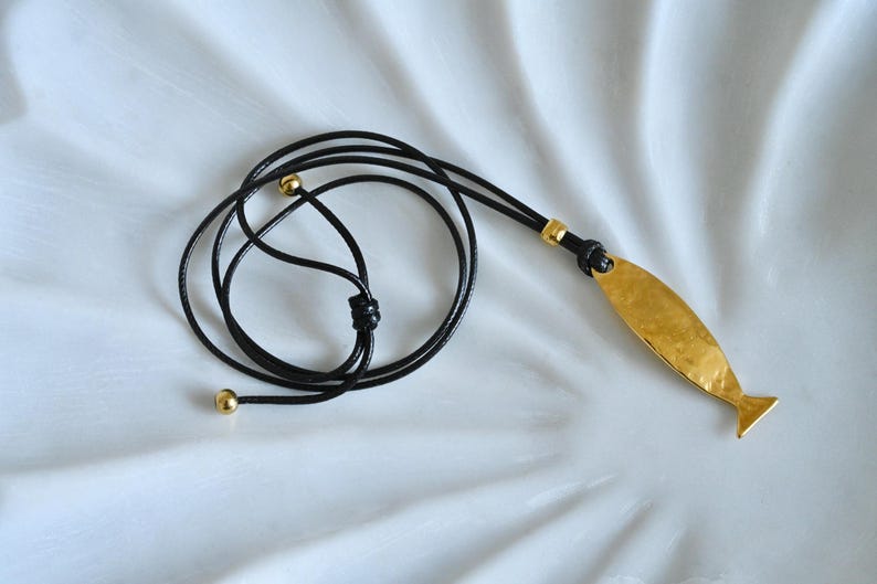 May include: A gold-colored pendant necklace with a fish-shaped charm. The pendant is suspended from a black cord with gold-colored accents. The necklace is displayed on a white surface.