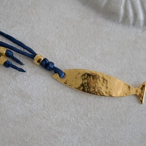 May include: A necklace featuring a gold-toned, hammered metal fish pendant attached to a dark blue cord. The cord includes three small, cylindrical gold-toned beads as decorative accents near the knot where the pendant is secured to the cord.
