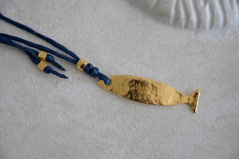 May include: A necklace featuring a gold-toned, hammered metal fish pendant attached to a dark blue cord. The cord includes three small, cylindrical gold-toned beads as decorative accents near the knot where the pendant is secured to the cord.