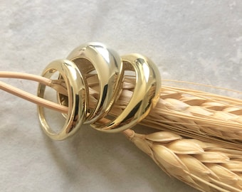 May include: Four gold-colored rings of varying sizes are displayed on a light-colored surface. The rings have a smooth, polished finish and a rounded design. A few pieces of wheat are in the background.