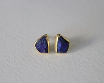 Può includere: Un paio di orecchini a lobo placcati in oro, ciascuno con una vibrante pietra di lapislazzuli blu scuro di forma irregolare. Gli orecchini sono posti su uno sfondo bianco sporco, evidenziando il contrasto tra l'oro e la gemma blu.