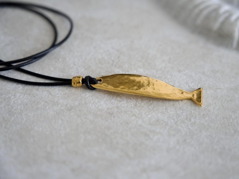May include: A gold-toned pendant shaped like a stylized fish with a hammered, textured surface. The pendant is attached to a thin black cord necklace by a small gold-toned bead and a knot, creating a simple and clean jewelry piece.