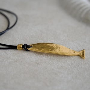 May include: A gold-toned metal pendant shaped like a stylized fish with a hammered, textured surface. The pendant is attached to a thin black cord necklace, which features a small gold-toned bead accent near the point of connection to the fish.