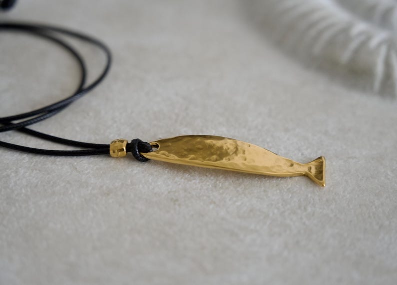 May include: A gold-toned metal pendant shaped like a stylized fish with a hammered, textured surface. The pendant is attached to a thin black cord necklace, which features a small gold-toned bead accent near the point of connection to the fish.