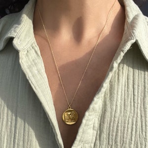 Op de afbeelding: Een goudkleurige ketting met een cirkelvormige hanger met een gedetailleerd reli&euml;fontwerp. De hanger hangt aan een fijne ketting, wat een klassieke en elegante uitstraling cre&euml;ert. De ketting wordt gedragen over een lichtgroen overhemd.