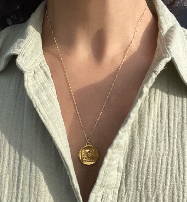 Op de afbeelding: Een goudkleurige ketting met een cirkelvormige hanger met een gedetailleerd reli&euml;fontwerp. De hanger hangt aan een fijne ketting, wat een klassieke en elegante uitstraling cre&euml;ert. De ketting wordt gedragen over een lichtgroen overhemd.