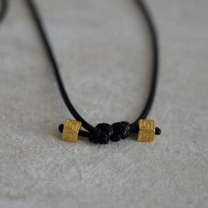 May include: A black cord necklace featuring two small, square, gold-toned metal beads with an etched geometric pattern. The beads are positioned on either side of a central black cord knot, creating a simple and symmetrical design element.