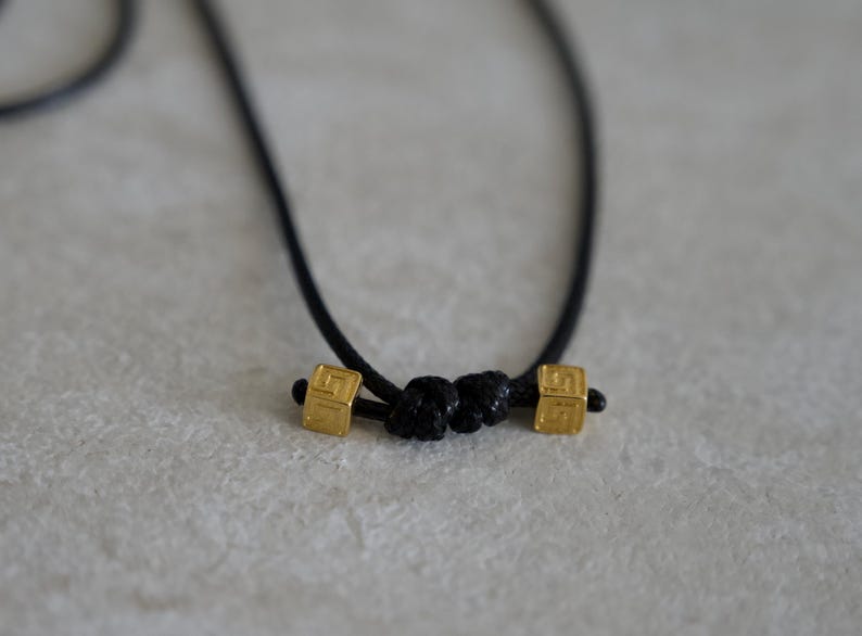 May include: A black cord necklace featuring two small, square, gold-toned metal beads with an etched geometric pattern. The beads are positioned on either side of a central black cord knot, creating a simple and symmetrical design element.