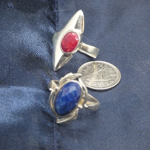 May include: Two silver rings with gemstone settings. The ring on the left has a blue gemstone and the ring on the right has a red gemstone. A Canadian coin is in the background.