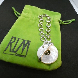 May include: A silver chain necklace with a large white mother-of-pearl pendant. The pendant is round and has a silver ring around the edge. The necklace is in a green velvet pouch with the text "RLM Studio" printed on it.