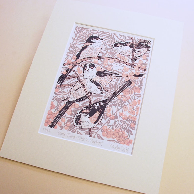 Op de afbeelding: Een linosnede van langstaartmezen die op een tak zitten met bessen. De vogels zijn afgebeeld in tinten roze en grijs tegen een achtergrond van roze en grijze bessen en bladeren. De print is getiteld "Long-tailed Tits in Berries" en gesigneerd "Eleanor Bell".