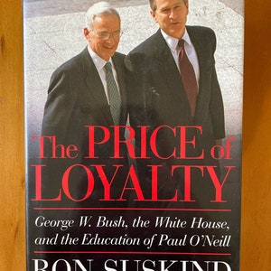 Pode incluir: Uma capa de livro com fundo preto e texto vermelho que diz "The Price of Loyalty". O subtítulo é "George W. Bush, the White House, and the Education of Paul O'Neill". O nome do autor, "Ron Suskind", está em texto branco abaixo do subtítulo. A capa do livro também apresenta uma foto em preto e branco de dois homens de terno.