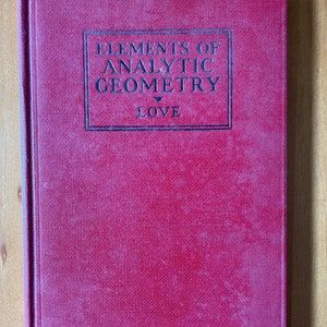 May include: A hardcover book with a textured, deep pink cover. The title "ELEMENTS OF ANALYTIC GEOMETRY" is printed in black within a rectangular frame, with the word "LOVE" below. The book is closed and resting on a wooden surface.