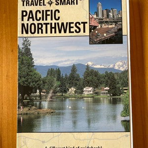 May include: A travel guide book cover with the title "TRAVEL SMART® PACIFIC NORTHWEST". The cover features a photo of a lake with mountains in the background and a city skyline in the upper right corner.