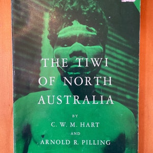 Op de afbeelding: Boekomslag met een groene achtergrond en een zwart-witfoto van een persoon met een geschilderd gezicht. De titel van het boek is "The Tiwi of North Australia" door C. W. M. Hart en Arnold R. Pilling.