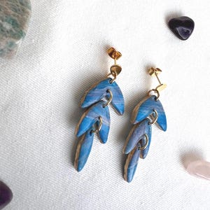 May include: Handmade earrings with a leaf design. Each earring has three blue and white marbled leaf-shaped pendants connected by gold-coloured hoops and posts. The earrings are displayed on a white surface with gemstones.