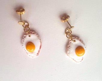 Könnte beinhalten: Ein Paar goldfarbene Ohrstecker mit Spiegelei-Design. Jeder Ohrring hat ein weißes Eiweiß mit einem gelben Eigelb und einen goldfarbenen Stecker und eine Kette.
