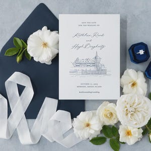 May include: A wedding invitation suite featuring a navy blue envelope, white ribbon, and a card with text and a line drawing of a building. White roses and two blue velvet ring boxes are also present.