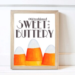 May include: A framed print featuring the words "Old fashioned SWEET n BUTTERY" in a decorative font. Below the text are three watercolor candy corn images with white, orange, and yellow colors. The frame is made of light-colored wood.