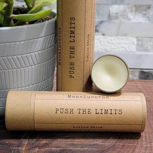 May include: Two brown cardboard tubes with the text "Manalucerna PUSH THE LIMITS Lotion Stick" printed on them. The tubes are stacked on top of each other on a wooden surface.