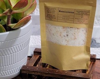 May include: A brown paper bag with a white label that reads "Manalucenna Relaxing Bath Salts 2 oz." The bag contains white bath salts with a hint of pink. The bag is sitting on a wooden surface.
