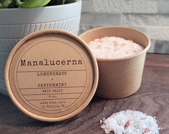 May include: A brown paper container of bath salts labeled "Manalucerna Lemongrass + Peppermint Bath Salts 4 oz. made with love in Dublin, VA"
