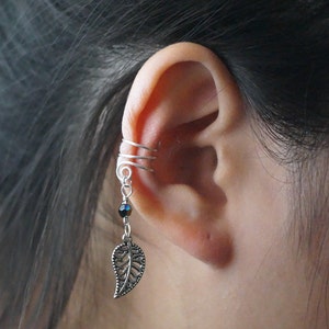 May include: Silver ear cuff with a dangling leaf charm and a black bead. The cuff is wrapped around the upper part of the ear.