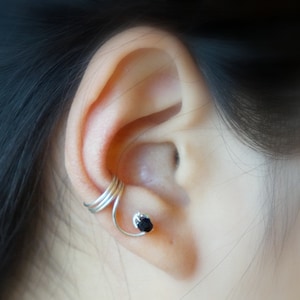 May include: Silver ear cuff with a black gemstone. The cuff wraps around the upper part of the ear and has a delicate design.
