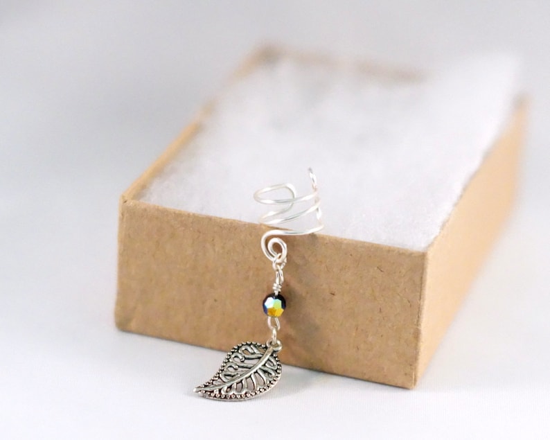 May include: Silver ear cuff with a spiral design and a dangling leaf charm. The charm is attached to a silver chain with a single iridescent bead.