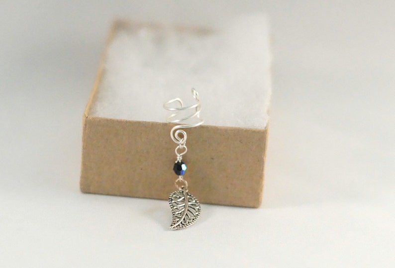 May include: Silver ear cuff with a spiral design and a dangling leaf charm. A black bead hangs above the leaf.