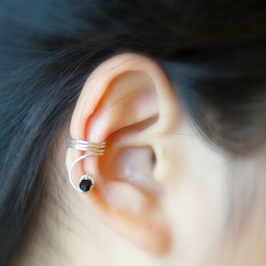 May include: A silver ear cuff with a black gemstone. The cuff is wrapped around the upper part of the ear.