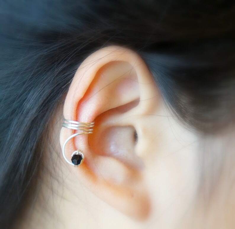 May include: A silver ear cuff with a black gemstone. The cuff is wrapped around the upper part of the ear.