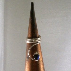 May include: A silver wire ring with a blue iridescent gemstone. The ring is displayed on a brown cone-shaped ring holder.