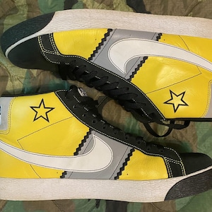 May include: Pair of high-top sneakers with yellow, black, gray, and white color-blocking. Features a white Nike swoosh, black laces, and a star graphic on the side. The sole is white with a black toe cap.