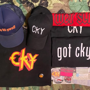 May include: A collection of CKY merchandise, including a navy blue trucker hat with orange text, a black baseball cap with white embroidery, and several black t-shirts with white text. Also included are stickers and tickets.