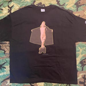 Könnte beinhalten: Schwarzes T-Shirt mit einem grafischen Aufdruck einer nackten Figur mit Umhang. Die Figur ist in Beige- und Brauntönen dargestellt. Das T-Shirt hat einen Rundhalsausschnitt und kurze Ärmel. Das Design ist mittig auf der Vorderseite.