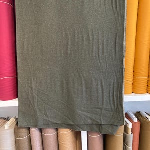 May include: A large piece of olive green fabric draped over a shelf, with rolls of various colored fabrics in the background. The fabric has a textured appearance. Other fabric rolls are red and yellow.