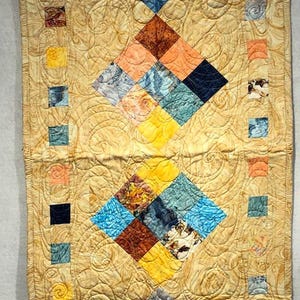 May include: A rectangular quilted table runner with a golden-yellow background and diamond-shaped patterns. The diamonds are composed of smaller squares in various colors, including blue, orange, brown, and yellow. The runner is likely handmade.