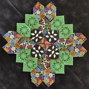 May include: A colorful, handmade quilt block with a geometric design. The block features green, black, and patterned fabric with circles and floral motifs. The quilt block is arranged in a symmetrical, star-like pattern.