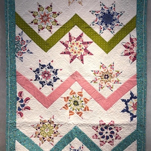 May include: A handmade quilt with a white background, featuring colorful star patterns and zigzag designs in green, pink, and teal. The quilt has a teal border with white polka dots and intricate stitching.