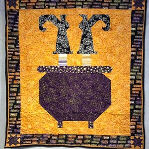 May include: A Halloween-themed quilt featuring a cauldron with witch legs and hats. The cauldron is purple with a spiderweb design, and the witch legs have striped socks. The background is orange, and the border has a brick pattern.