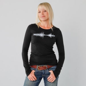 Puede incluir: Una camiseta negra de manga larga con un gráfico de onda sonora blanca en la parte delantera. La camiseta es entallada y tiene un cuello redondo.