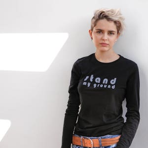 Puede incluir: Una mujer con el pelo corto y rubio lleva una camiseta negra de manga larga con el texto "stand my ground" impreso en la parte delantera. Lleva un cinturón de cuero marrón con una hebilla plateada.