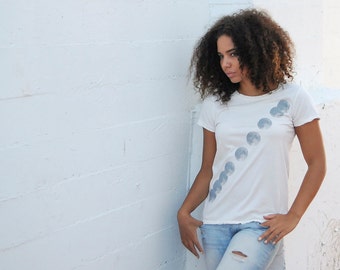 May include: A white t-shirt with a graphic of a crescent moon in various phases printed on the front. The shirt is worn by a person with curly brown hair against a white wall.