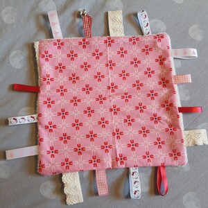 May include: A pink and white baby sensory blanket with various textures and ribbons. The blanket features a pattern of safety pins and has a white, fluffy border. The ribbons are in various colors, including red, white, blue, and pink.